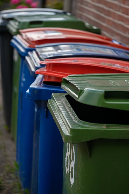 A row of seven wheelie bins lined up along a pavement beside a brick wall, each with a distinct colour lid, including green, blue, red, and dark green. The green bin in the foreground has a textured,