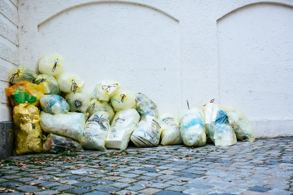 A row of seven wheelie bins lined up along a pavement beside a brick wall, each with a distinct colour lid, including green, blue, red, and dark green. The green bin in the foreground has a textured,