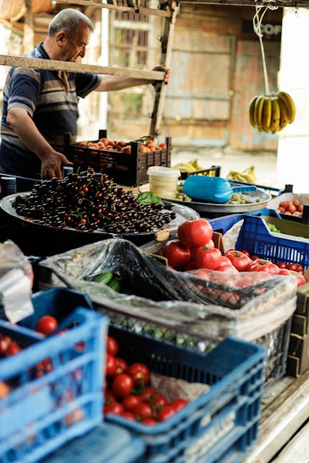 The image depicts a man at an outdoor market stall preparing fresh produce, with various crates and containers filled with tomatoes, cherries, and green vegetables arranged on a wooden table or counte
