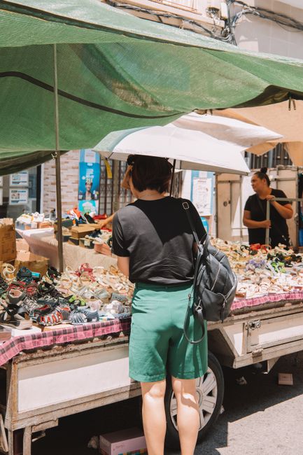 The image depicts a man at an outdoor market stall preparing fresh produce, with various crates and containers filled with tomatoes, cherries, and green vegetables arranged on a wooden table or counte